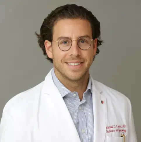 Dr. Michael Lewis, a young man with glasses and brown hair wearing a button down shirt and physician's lab coat.